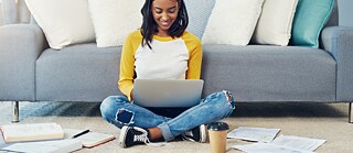 Eine junge Frau lernt online am Laptop. Sie sitzt gemütlich auf dem Boden im Schneidersitz. Sie lehnt an ein Sofa. Auf dem Boden liegen verstreut Bücher und lose Arbeitsblätter. Vor ihr steht ein Pappbecher mit einem Kaffeegetränk.